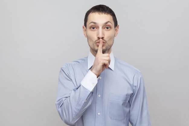 Please be quiet. Portrait of serious handsome bristle businessman in classic light blue shirt standing and looking at camera with silent sign. indoor studio shot, isolated on grey background copyspace