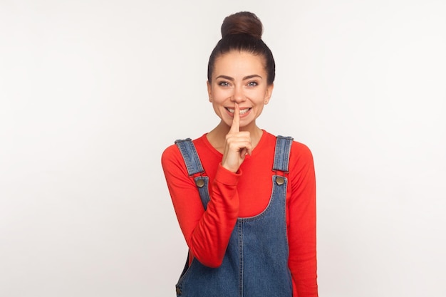 Please don39t talk Portrait of positive stylish pretty girl with hair bun in denim overalls shushing asking for silence or secrecy with finger on lips be quiet indoor studio shot white background