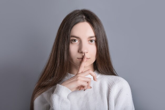 Foto per favore, non parlare. misteriosa donna dai capelli scuri chiede di tacere, preme l'indice sulle labbra, esige il silenzio, racconta informazioni riservate segrete, isolatad su sfondo grigio, dice zitto.