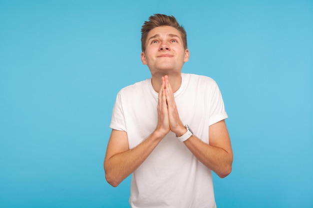 Please, God I'm begging. Portrait of beseeching man in casual white t-shirt looking up and praying, pleading with sorrowful imploring expression. indoor studio shot isolated on blue background