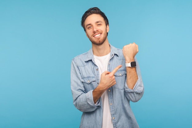 Per favore, faccia in fretta. ritratto di uomo allegro e puntuale con una camicia di jeans da lavoro che punta all'orologio da polso e sorridente, mostrando lo smartwatch con display mock up. indoor isolato su sfondo blu