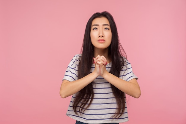 お願いします、Tシャツを着た長い髪の心配している女の子の肖像画を見上げて、懇願するような目で祈って、助けを求めて、許しを求めます。ピンクの背景に分離されたスタジオショット