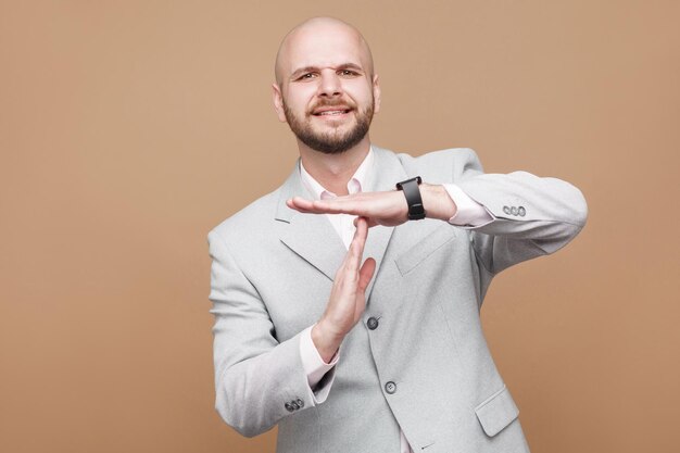 Per favore, ho bisogno di più tempo. lavoratore d'affari barbuto calvo di mezza età in classico abito grigio chiaro in piedi guardando la fotocamera e chiedendo il timeout. studio indoor girato, isolato su sfondo marrone.