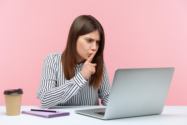 Пожалуйста, держите в секрете. Обеспокоенная женщина-сотрудник, сидящая на рабочем месте и молчащая жестом, разговаривает по видеозвонку, онлайн-общение через ноутбук. Снимок в закрытой студии изолирован на розовом фоне.