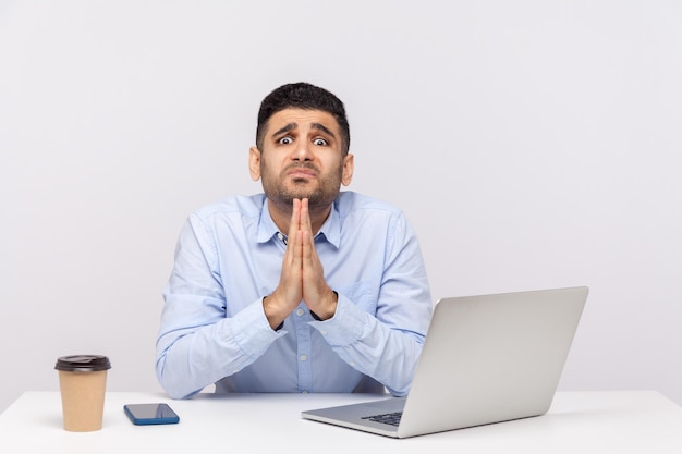 Please need help Upset man employee sitting office workplace with laptop on desk holding prayer gesture asking heartily pleading with desperate grimace studio shot isolated on white background