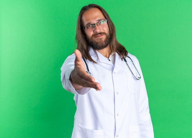 Pleased adult male doctor wearing medical robe and stethoscope with glasses looking at camera doing greeting gesture isolated on green wall with copy space