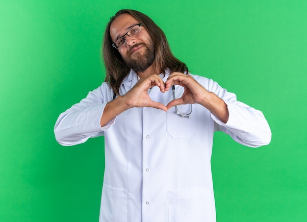 Pleased adult male doctor wearing medical robe and stethoscope with glasses looking at camera doing love sign isolated on green wall