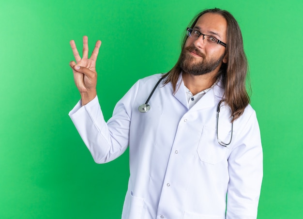 Pleased adult male doctor wearing medical robe and stethoscope with glasses looking at camera showing three with hand isolated on green wall