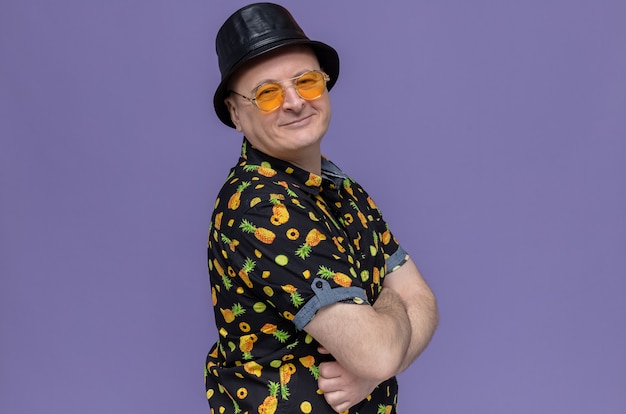 Pleased adult man with black top hat wearing sunglasses standing sideways with crossed arms and looking 