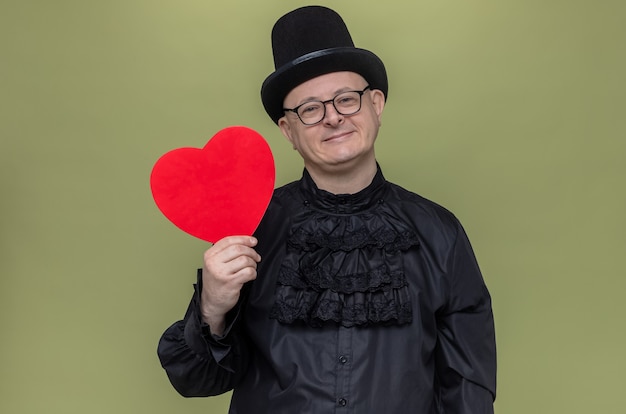 Pleased adult slavic man with top hat and optical glasses in black gothic shirt holding red heart shape and looking at front