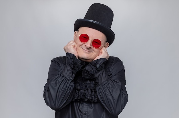 Pleased adult slavic man with top hat and with sunglasses in black gothic shirt putting fists on his face and looking at front