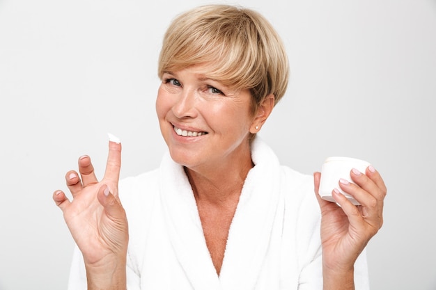 Compiaciuta donna adulta con corti capelli biondi che indossa una vestaglia bianca che tiene un vasetto di crema per il viso isolato su un muro bianco