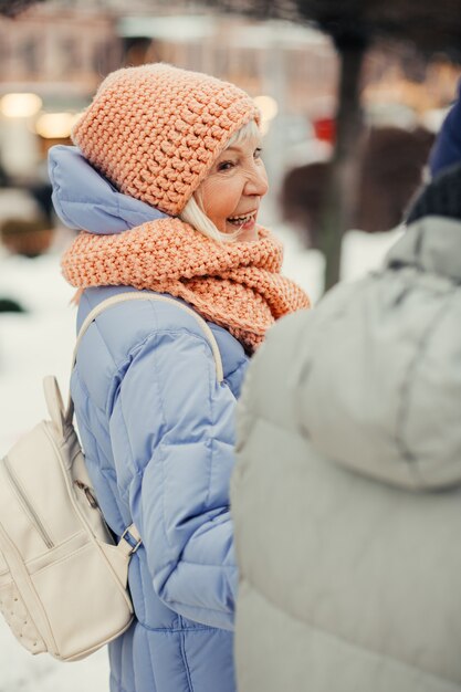 Pleased aged lady in winter clothes with backpack laughing and looking at the person next to her