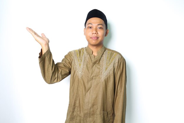 白い背景に開いた手のひらで横に提示するココの服を着て満足しているアジアのイスラム教徒の男性