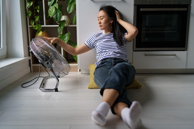 Pleased Asian woman blown by electric fan sits on floor enjoy cool wind from air ventilator in heat