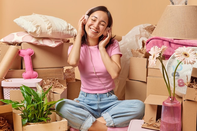 Pleased asian woman enjoys listening music in stereo headphones\
sits crossed legs at messy room relocates to new apartment buys new\
house for living happy property owner surrounded by cardboard\
boxes