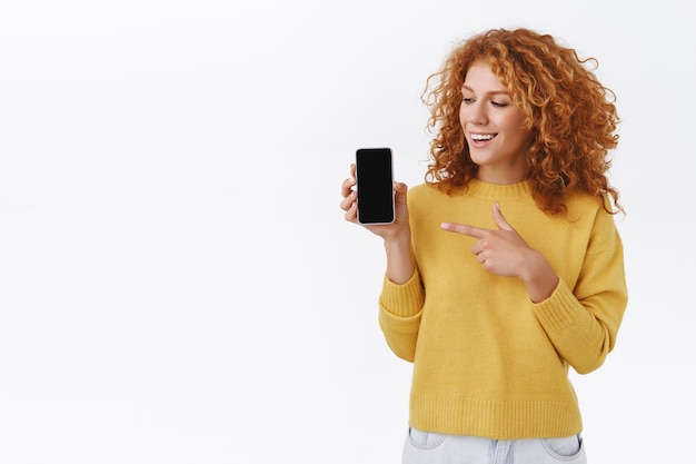 Piacere attraente donna riccia rossa in maglione giallo, tenendo in mano lo smartphone, indicando il display mobile e sorridendo, consiglia l'applicazione, muro bianco