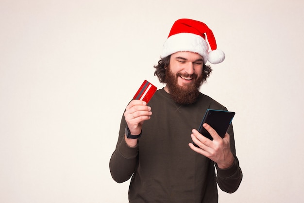 Pleased bearded man is using his bank card, ordering online and looking at his tablet.