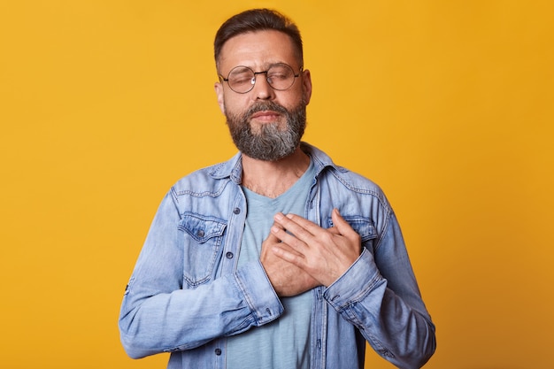 Pleased bearded man with closed eyes, keeps hands on chest, dressed in fashionable denim jacket