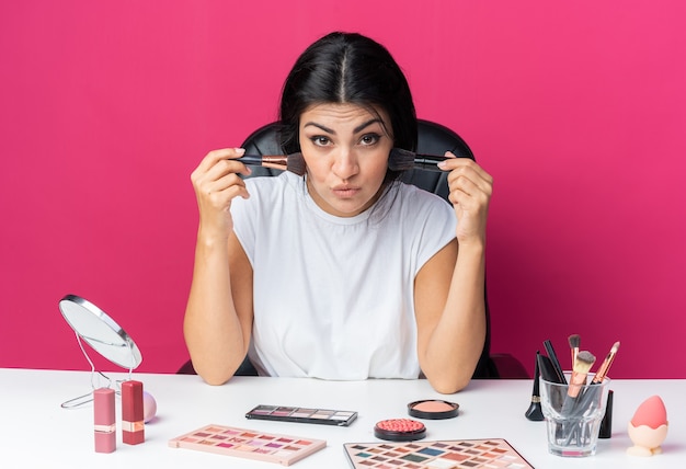 Pleased beautiful woman sits at table with makeup tools holding powder brushes around face 