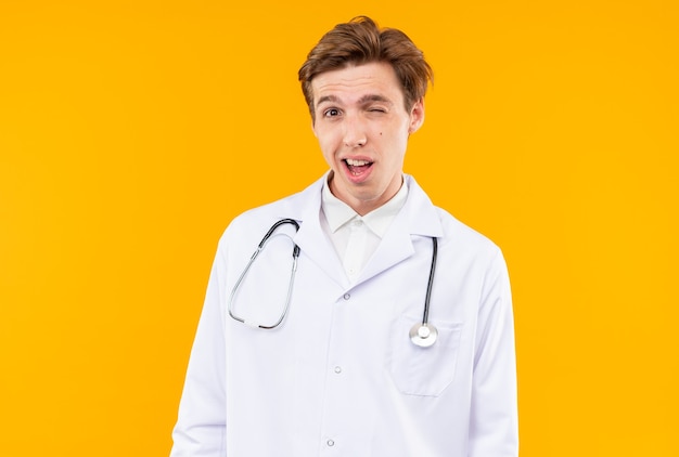 Pleased blinked young male doctor wearing medical robe with stethoscope isolated on orange wall