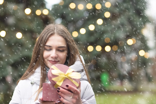 Pleased blond woman holding a gift box at the street during the snowfall. Empty space