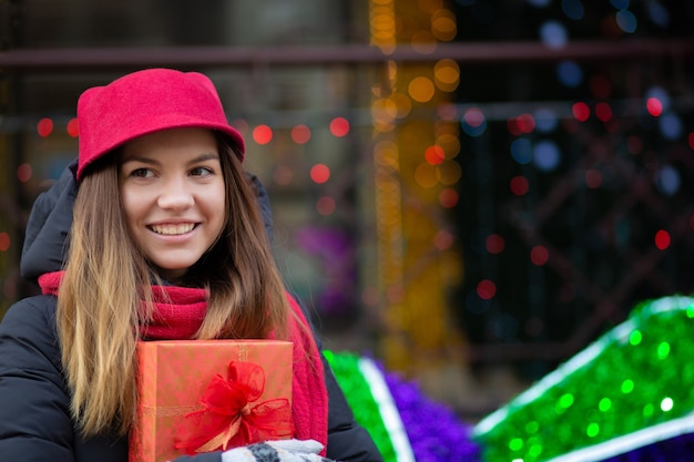 Pleased blonde woman in red hat and knit scarf holding gift box. Space for text