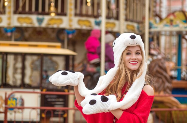 Pleased blonde woman wearing red knitted sweater and funny hat, posing at the background of carousel with lights