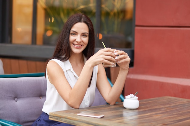 Pleased brunette lady holds fresh cocktail, sits at wooden table in cozy outdoor cafeteria, uses modern mobile phone