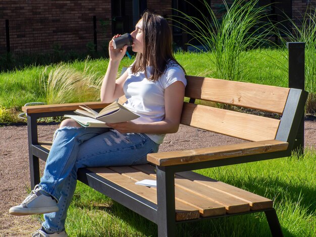 ベンチに座って公園でコーヒーを飲む満足したブルネットの女性 教育学校と人々のコンセプト キャンパスのベンチで勉強する女子学生 本のコーヒーカップで座って書いている女の子