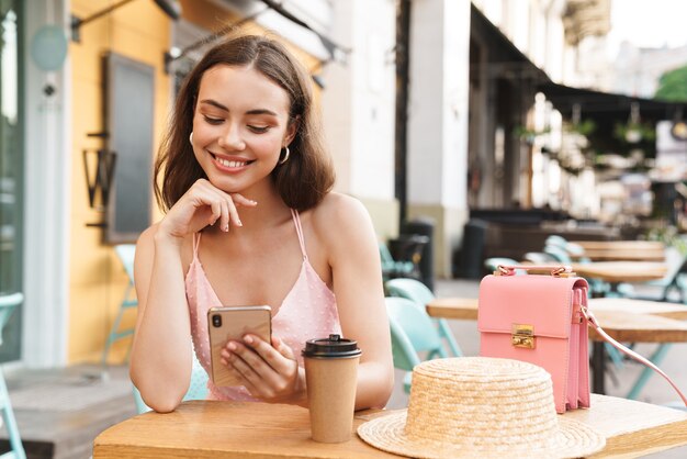 pleased brunette woman smiling and holding cellphone while sitting in street summer cafe with takeaway coffee