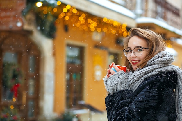 Pleased brunette woman wears knit scarf and stylish glasses enjoying snowfall and drinks coffee. Empty space