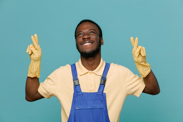 Pleased crossing fingers young africanamerican cleaner male in uniform with gloves isolated on blue background