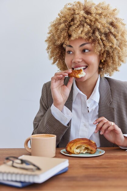 Pleased curly haired successful woman bites delicious croissant\
drinks coffee dressed in formal outfit poses at woodent table with\
notepads and spectacles on them has delicious snack in\
cafeteria