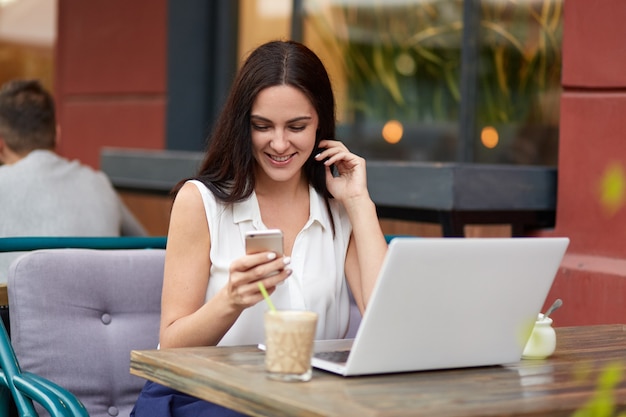 Pleased dark haired woman holds modern cell phone, reads positive news on internet website, drinks cocktail in outdoor cafeteria