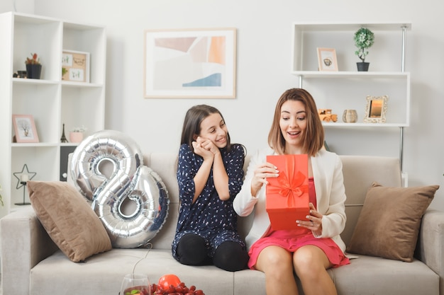 Pleased daughter gives present to surprised mother on happy woman's day sitting on sofa in living room