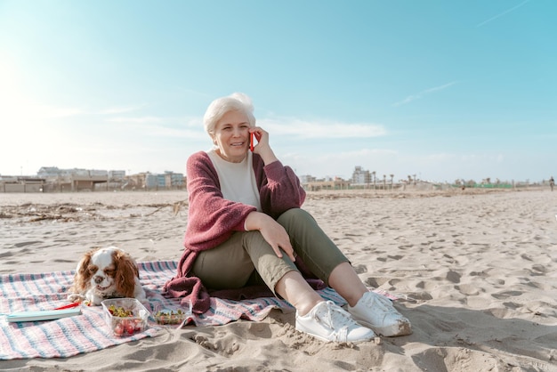 Piacevole signora anziana seduta sulla sabbia accanto al suo simpatico animale domestico durante il colloquio telefonico