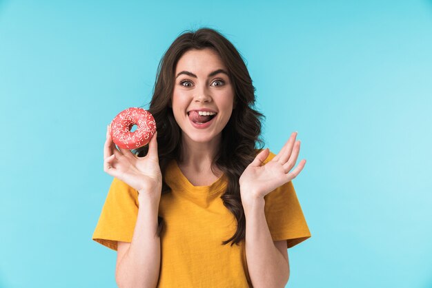 기쁘게 흥분된 긍정적인 젊은 예쁜 여자 도넛을 들고 파란색 벽 위에 고립 된 포즈.