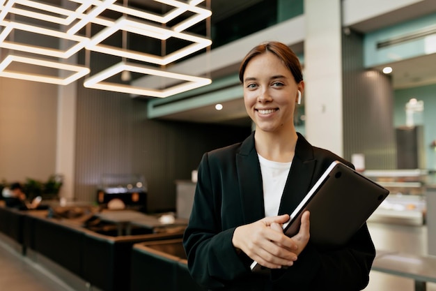 Pleased female in trendy jacket posing in office after meeting with colleagues Indoor portrait of stylish businesswoman is holding laptop in headphones