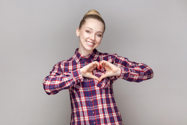 Pleased friendly woman looking at camera with charming smile showing heart shape gesture