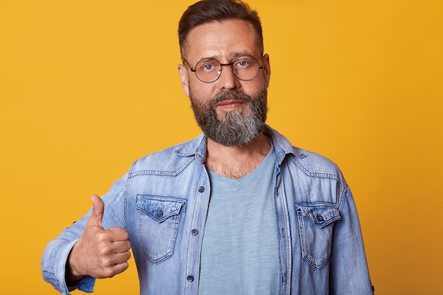 Pleased and good looking male in sunglasses, denim jacket and gray t shirt, raising thumb up and