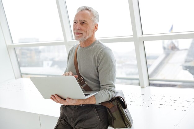 pleased gray-haired man holding and using laptop while standing near windows indoors