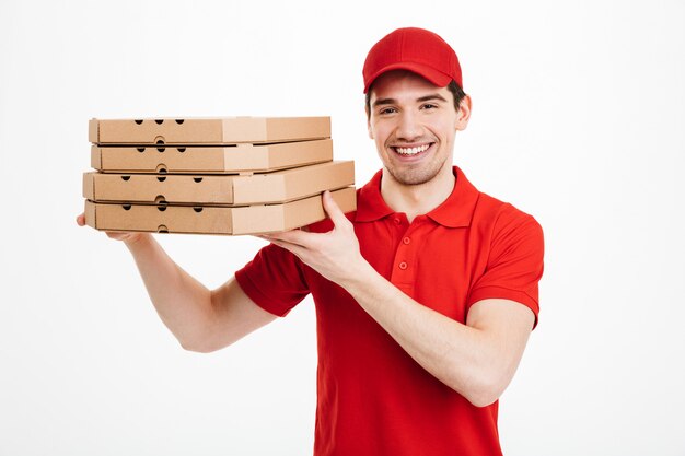 Pleased guy dealer in red t-shirt and cap working in delivery service and holding stack of pizza boxes, isolated over white space