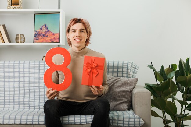 Pleased handsome guy on happy women day holding number eight with present sitting on sofa in living room