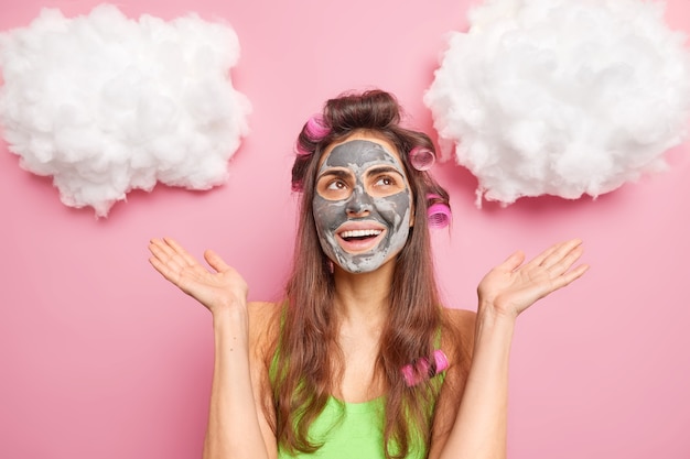 Pleased happy Caucasian woman applies beauty mask on face spreads palms and looks happily above prepares for date wants to have fabulous look isolated over pink wall