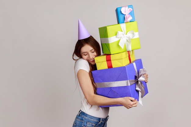 Foto donna felice contenta che abbraccia molti regali di compleanno e sorride con gioia, festeggia, ammira molti regali, indossa una maglietta bianca. studio indoor girato isolato su sfondo grigio.