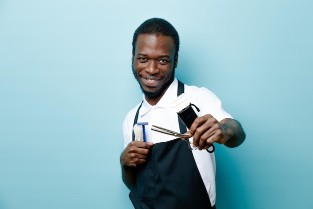 Pleased holding barber tools young african american barber in uniform isolated on blue background