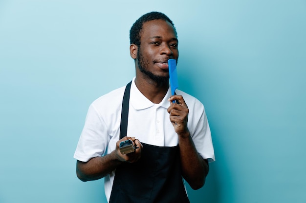 Pleased holding comb with hair clipper young african american barber in uniform isolated on blue background