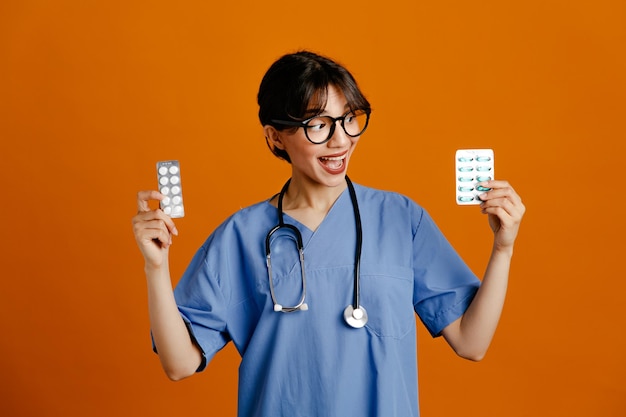 Felice azienda pillole giovane dottoressa che indossa uno stetoscopio fith uniforme isolato su sfondo arancione