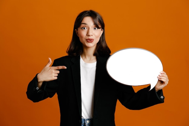 Pleased holding and points at speech bubble young beautiful female wearing black jacket isolated on orange background